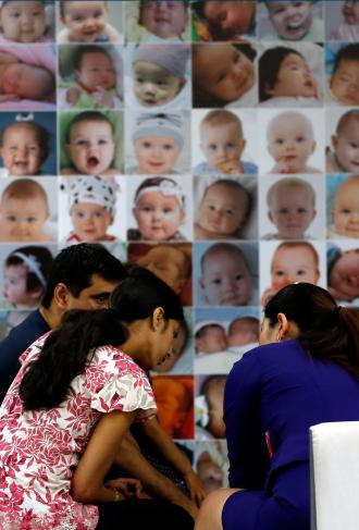 Tre voksne i samtale foran vegg dekket av babyportretter (samme bilde som over)